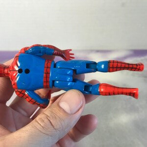 May include: A red and blue plastic action figure of Spider-Man. The figure is posed with its arms outstretched and legs bent. The figure has a black dot on its back.
