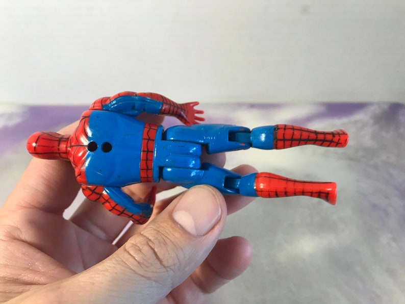May include: A red and blue plastic action figure of Spider-Man. The figure is posed with its arms outstretched and legs bent. The figure has a black dot on its back.