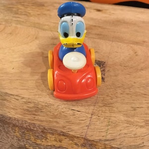 May include: A vintage toy car with a yellow, orange and white design. A cartoon duck figure is sitting in the driver's seat.