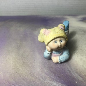 May include: A porcelain figurine of a girl with blonde hair, blue eyes, and a pink and blue dress. She is sitting with her chin resting on her hand.