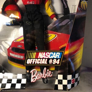 May include: A Barbie doll wearing a red and yellow racing suit with the words "Drive Thru" and "Coca-Cola" on the front. The doll is holding a red helmet and standing in front of a red race car with the words "Taurus" and "NASCAR Official #94" on the side. The box also has the words "Barbie" and "McDonald's" on it.