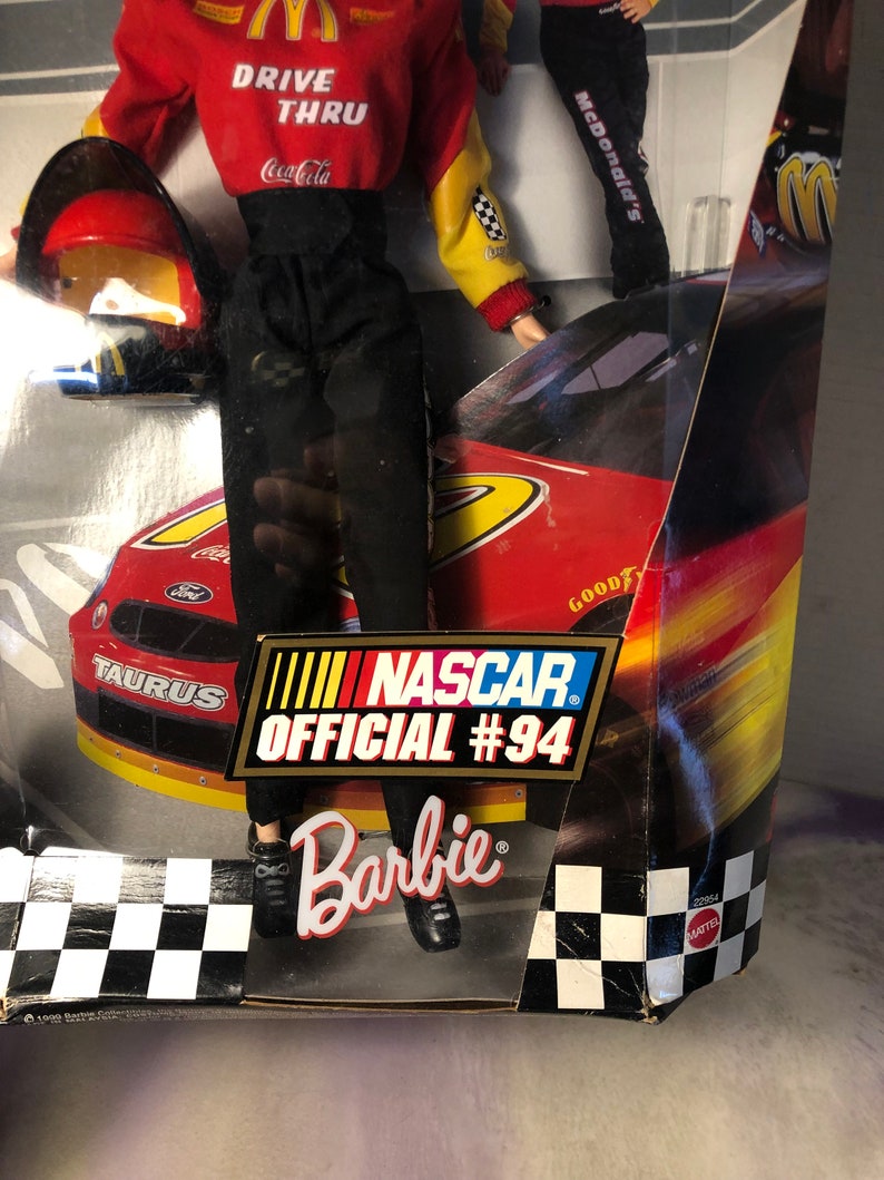 May include: A Barbie doll wearing a red and yellow racing suit with the words "Drive Thru" and "Coca-Cola" on the front. The doll is holding a red helmet and standing in front of a red race car with the words "Taurus" and "NASCAR Official #94" on the side. The box also has the words "Barbie" and "McDonald's" on it.