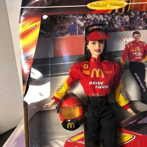 May include: A red, yellow, and black NASCAR racing doll with a red helmet and a McDonald's Drive Thru uniform. The doll is holding a Coca-Cola can. The doll is standing in front of a red race car with the word "TAURUS" on the side.