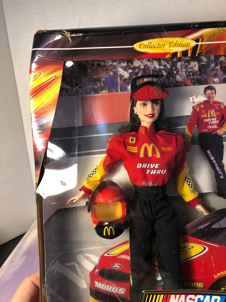 May include: A red, yellow, and black NASCAR racing doll with a red helmet and a McDonald's Drive Thru uniform. The doll is holding a Coca-Cola can. The doll is standing in front of a red race car with the word "TAURUS" on the side.