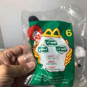 May include: A plastic bag containing a white plush toy of Goofy from Disney's House of Mouse. The bag is green and features the McDonald's logo and the text "House of Mouse" and "Mickey Mania".