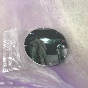 Può includere: Un oggetto da collezione rotondo di Star Wars color argento che presenta una scena del film con Luke Skywalker e la Principessa Leia in piedi davanti a R2-D2.