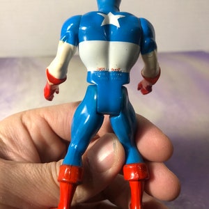 May include: A vintage Captain America action figure, viewed from the back. The figure is primarily blue with white and red accents. The back features a white star on a blue field. The figure is held in a hand.