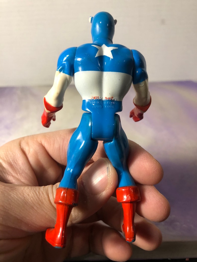May include: A vintage Captain America action figure, viewed from the back. The figure is primarily blue with white and red accents. The back features a white star on a blue field. The figure is held in a hand.
