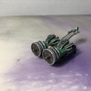 以下が含まれることがあります： 緑と銀色のモデルの宇宙船。2つの大きなエンジンと細長い胴体を持つ。宇宙船は白と紫の表面の上に置かれている