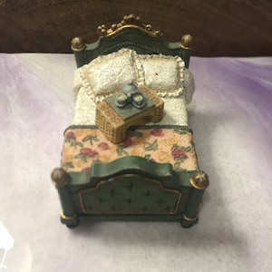 May include: A miniature green bed with a gold trim and a floral patterned bedspread. A small wooden table with a tea set sits on the bed.