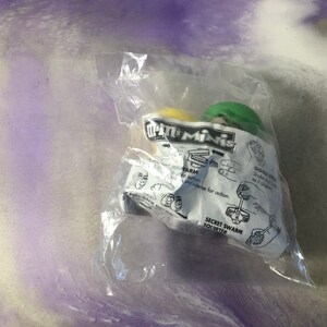 Könnte beinhalten: Eine Plastiktüte mit zwei kleinen grünen und gelben M&M's Minis Spielzeugen. Die Verpackung zeigt das M&M's Minis Logo und Anweisungen zur Verwendung der Spielzeuge. Die Spielzeuge sind mit "Secret Swarm Squirter" gekennzeichnet.