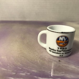 Może przedstawiać: Mały biały ceramiczny kubek z czarnym obramowaniem i uchwytem. Kubek zawiera logo New York Islanders i tekst upamiętniający ich zwycięstwo w Pucharze Stanleya w 1983 roku, w tym wynik finałowego meczu.