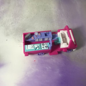 Op de afbeelding: Een roze en paarse speelgoed foodtruck met een keuken en een zithoek. De truck heeft een wit dak en een blauw en wit interieur. De keuken heeft een gootsteen, een fornuis en andere keukenaccessoires.