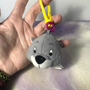 May include: A grey plush seal keyring with a yellow clip and purple detail. The seal has a black nose, white eyes, and white accents. The keyring is attached to a yellow plastic clip.