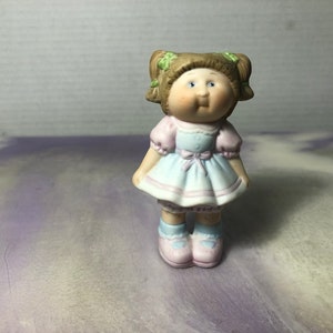 May include: A porcelain figurine of a little girl with brown hair in pigtails, wearing a light blue dress with a pink bow and pink shoes.