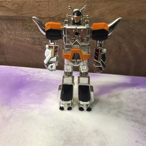 May include: A silver and orange robot toy with articulated arms and legs. The robot has a black head and accents, with silver wings extending from its shoulders. The toy is standing on a white and purple surface.
