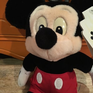 Może przedstawiać: Pluszowa zabawka Mickey Mouse w kolorze czarnym i białym, ubrana w czerwone spodenki z białymi k&oacute;łkami. Zabawka ma r&oacute;żową twarz i duże czarne oczy.