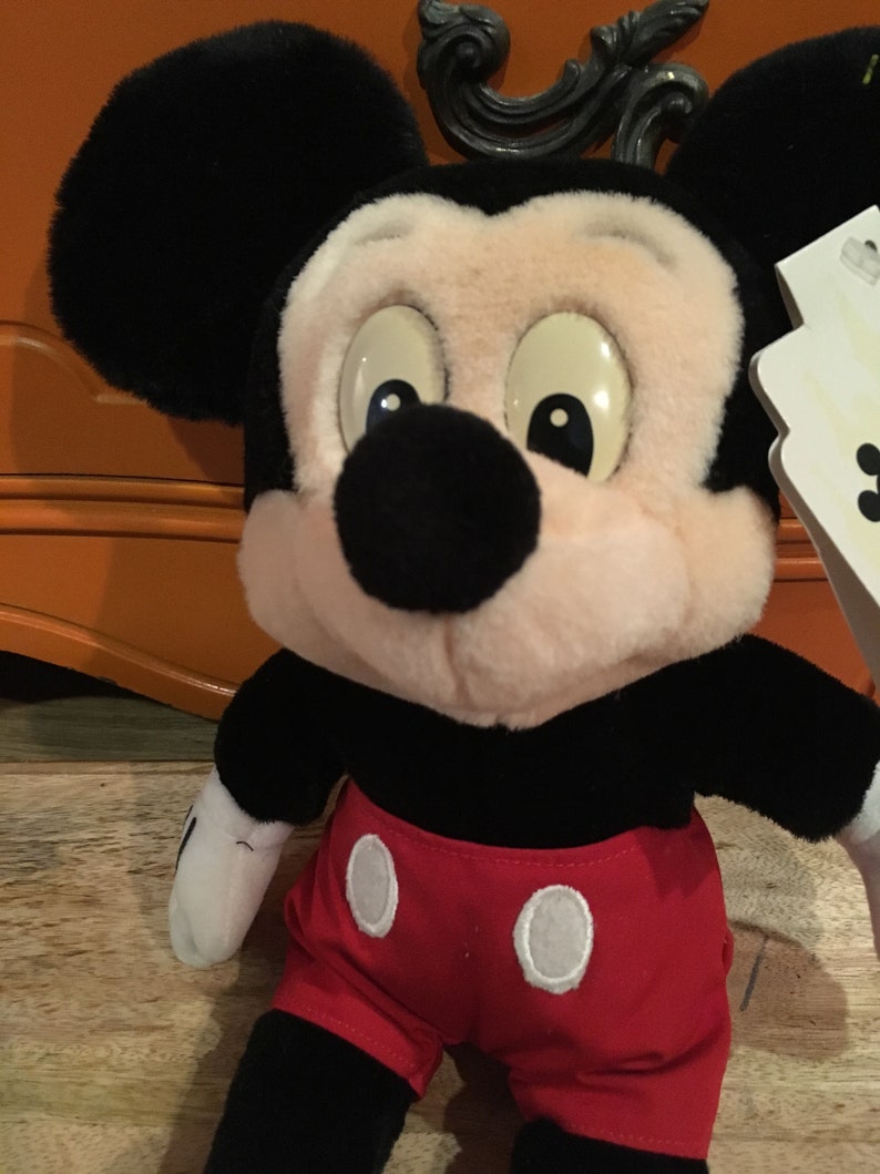 Może przedstawiać: Pluszowa zabawka Mickey Mouse w kolorze czarnym i białym, ubrana w czerwone spodenki z białymi k&oacute;łkami. Zabawka ma r&oacute;żową twarz i duże czarne oczy.