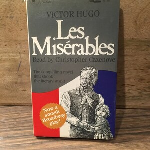 May include: A grey cassette tape case with the title "Les Misérables" by Victor Hugo. The case features a black and white illustration of a man and a young girl. The text "Read by Christopher Cazenove" is also on the case.