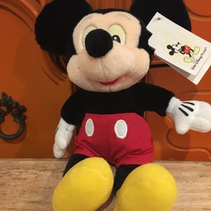 Puede incluir: Un juguete de peluche de Mickey Mouse sentado con una camisa roja y blanca y zapatos amarillos. El juguete tiene una nariz negra y orejas negras grandes. El juguete tiene una etiqueta blanca unida a su oreja derecha con el texto "Walt Disney World".
