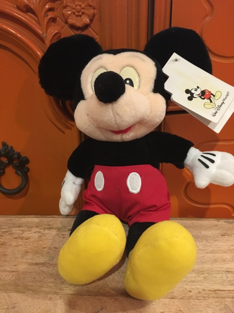 Puede incluir: Un juguete de peluche de Mickey Mouse sentado con una camisa roja y blanca y zapatos amarillos. El juguete tiene una nariz negra y orejas negras grandes. El juguete tiene una etiqueta blanca unida a su oreja derecha con el texto "Walt Disney World".