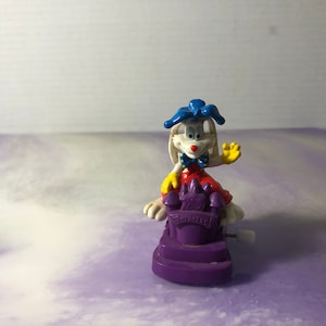 May include: A wind-up toy of Bugs Bunny wearing a red and white outfit and a blue hat, sitting on a purple base with a yellow "20th Anniversary" inscription.