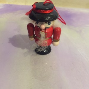 May include: A wooden nutcracker ornament with a red and black uniform, a white beard, and a black hat. The ornament has a red string attached to the top.