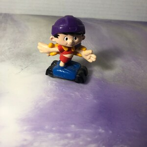 May include: A purple and yellow plastic toy figure of a boy wearing a purple helmet and riding a blue scooter.