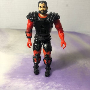 Könnte beinhalten: Eine schwarze und rote Actionfigur mit schwarzem Bart und schwarzer Rüstung. Die Figur hat einen ernsten Gesichtsausdruck.