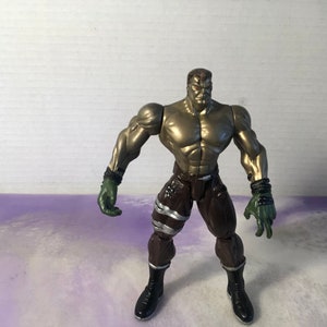 Può includere: Una action figure dorata con un fisico muscoloso, che indossa pantaloni marroni con dettagli argentati e guanti verdi. La figura ha un'espressione seria sul viso.
