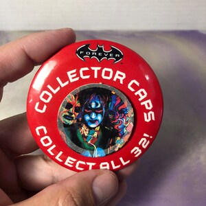 Può includere: Spilla rotonda rossa con il testo "Collector Caps! Collect All 32!" e un logo di Batman con la parola "Forever" sotto. La spilla presenta un'immagine colorata di un personaggio con un costume verde e nero e un viso blu.