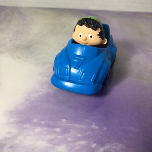 May include: A blue toy race car with a number 7 on the side. A small cartoon figure with black hair and a green hat is sitting in the driver's seat.