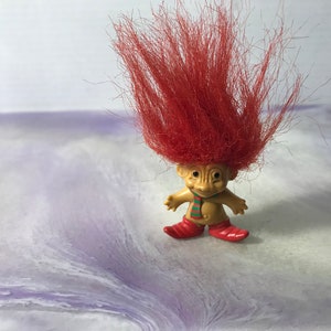 Può includere: Un piccolo troll di plastica con i capelli rossi, una sciarpa a righe verde e bianca e scarpe rosse. Il troll è in piedi su una superficie bianca e viola.