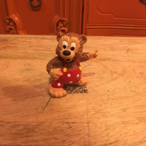 以下が含まれることがあります： 白い水玉模様の赤いオーバーオールを着た茶色のクマの置物。クマはオーバーオールに黄色のボタンが付いていて、口を開けて笑っています。置物は木製の表面に立っています。