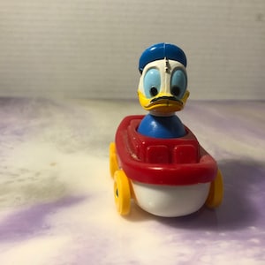 May include: A red and white toy boat with yellow wheels. A cartoon duck with a blue hat is sitting in the boat.