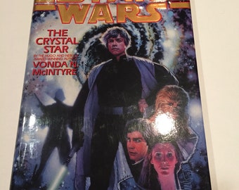 Może przedstawiać: Twarda oprawa książki zatytułowana "Star Wars: The Crystal Star" z okładką przedstawiającą Luke'a Skywalkera, Chewbaccę i inne postacie. Tytuł jest w złotych literach, a tło to scena kosmiczna.