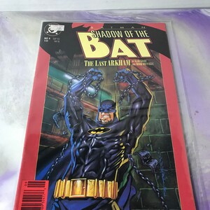 May include: A comic book cover featuring Batman in a dark blue and black suit, chained and struggling against a dark background. The title of the comic is "Batman: Shadow of the Bat" and the subtitle is "The Last Arkham".