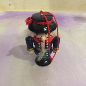 May include: A wooden nutcracker ornament with a black hat, blue jacket, and white beard. The ornament is decorated with red and white accents and has a red string attached to it.
