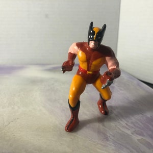 May include: A Wolverine action figure in a dynamic pose. The figure features a yellow and brown costume with black accents, including a mask and claws. The figure is approximately 4 inches tall.