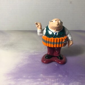 Może przedstawiać: Mała plastikowa figurka mężczyzny w zielonej kamizelce, czerwonych spodniach i białej koszuli. Ma wąsy i okulary i nosi pas z dynamitu wokół talii.