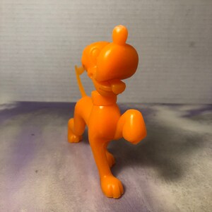 Peut inclure: Figurines de chien en plastique orange. Le chien est debout sur ses quatre pattes, la patte droite levée. Le chien a une longue queue et une grosse tête avec un visage simple.