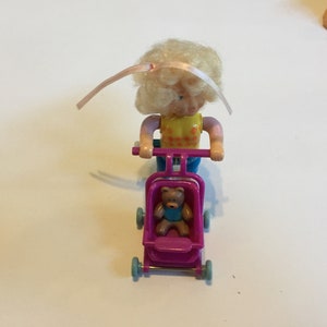 May include: A blonde doll with a pink ribbon in her hair pushes a pink stroller with a brown teddy bear inside. The stroller has blue wheels.