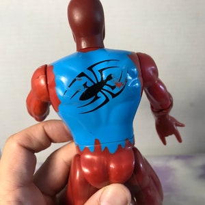 Puede incluir: Figura de acci&oacute;n roja y azul con un dise&ntilde;o de ara&ntilde;a negra en la espalda. La figura tiene una cabeza y un torso rojos, brazos y piernas azules, y un dise&ntilde;o de ara&ntilde;a negra en la espalda.