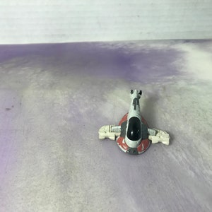 Pode incluir: Uma pequena nave espacial de brinquedo com um esquema de cores branco, vermelho e cinza. O brinquedo tem um cockpit preto e está sobre um fundo roxo claro e branco. A nave espacial tem asas e um design detalhado.