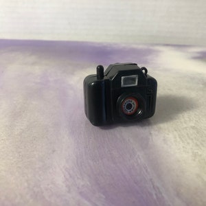 May include: A small black toy camera with a red circle lens and a silver flash. The camera has a black strap.