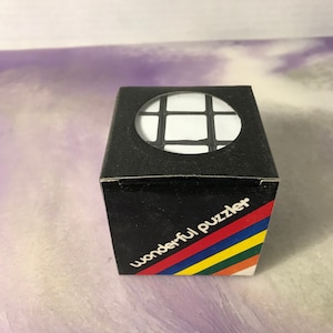 May include: A black box with a white cube inside. The box has a rainbow stripe design and the text "wonderful puzzler" on the front.