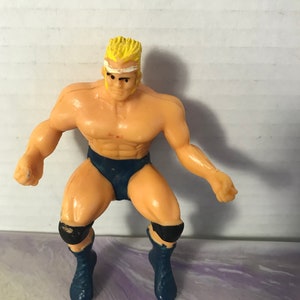 May include: A plastic action figure of a wrestler with blonde hair, a white headband, a blue wrestling outfit, and black knee pads.