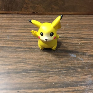 May include: A yellow Pikachu figurine with black ears and a white face. The figurine is standing on a wooden surface.