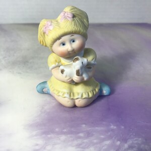 May include: A ceramic figurine of a seated child in a yellow dress, holding a small white and brown lamb. The child has blonde hair with pink bows and blue shoes. The figurine is on a purple and white surface.
