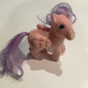 Könnte beinhalten: Ein rosa Plüschpony mit lilafarbenem Haar und Flügeln. Das Pony hat blaue Augen und ein glitzerndes Finish.